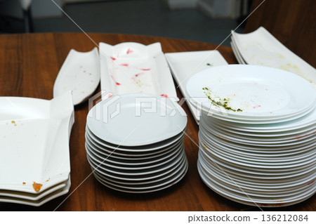 Stacked dirty plates and empty serving dishes after a banquet, showcasing leftover food remnants on a wooden table, emphasizing post-event cleanup and dining aftermath Stacked dirty plates and empty serving dishes after a banquet, showcasing leftover food remnants on a wooden table, emphasizing post-event cleanup and dining aftermath 136212084