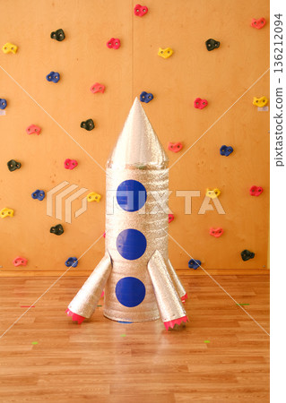 Homemade space rocket for children, crafted from shiny materials with colorful accents, standing on wooden floor against a playful climbing wall background, inspiring imaginative play and creativity Homemade space rocket for children, crafted from shiny materials with colorful accents, standing on wooden floor against a playful climbing wall background, inspiring imaginative play and creativity 136212094