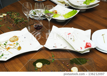 Elegant dining table with empty plates and wine glasses, showcasing remnants of a gourmet meal, surrounded by fresh greens and vibrant garnishes, creating a sophisticated culinary atmosphere 136212097