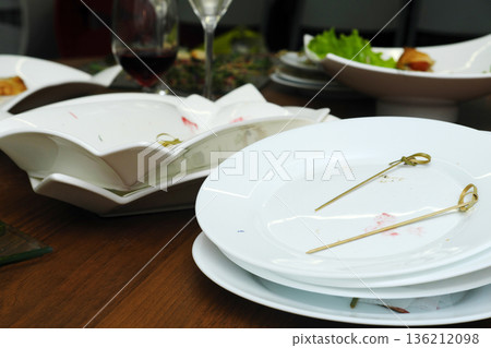Dirty dishes piled high on a wooden table after a banquet, featuring leftover food remnants and elegant serving platters, showcasing the aftermath of a festive reception 136212098