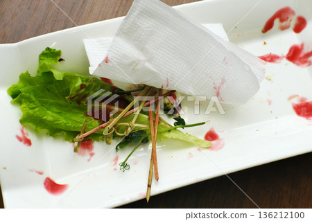Leftover food remnants on a white plate with dirty napkin, wilted lettuce, and sauce stains, depicting the aftermath of a dining event with unclean dishes 136212100