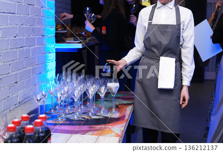 Professional catering service staff member in gray apron pouring drinks into elegant glasses at a stylish event, showcasing hospitality and attention to detail with copy space 136212137