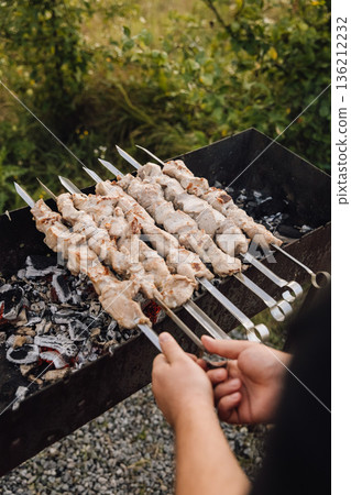 Hands turning pork shashlik skewers over charcoal grill, outdoor barbecue cooking 136212232