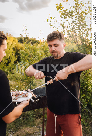 Man serving grilled pork shashlik from skewer to plate at outdoor barbecue Man serving grilled pork shashlik from skewer to plate at outdoor barbecue 136212244