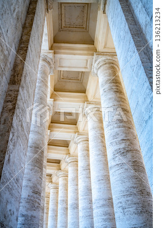 The view shows tall columns in a colonnade at St Peters Square in Vatican. The structure highlights classic architecture and historical design. Light comes through the columns creating a unique view. 136213234