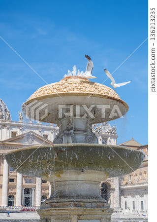 Water flows from the Bernini Fountain in St Peters Square. Birds stand on the fountain's edge. Visitors explore the square under a clear blue sky. The architectural details surround the area. Water flows from the Bernini Fountain in St Peters Square. Birds stand on the fountain's edge. Visitors explore the square under a clear blue sky. The architectural details surround the area. 136213235