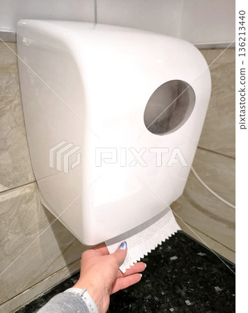 A woman's hand pulls out a white paper towel from a wall-mounted dispenser. 136213440