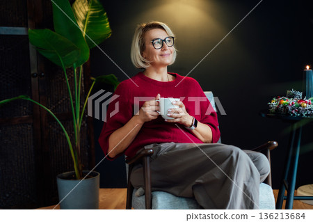 Middle aged woman relaxing with cup of hot drink in scandy style hygge interior home with winter mood decor. Lady dreaming, enjoy calm mood without stress, well being alone. Cozy fall winter holidays. 136213684