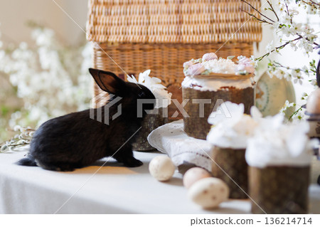 Black bunny with Easter cakes and eggs on spring table, blossom branches and wicker basket 136214714