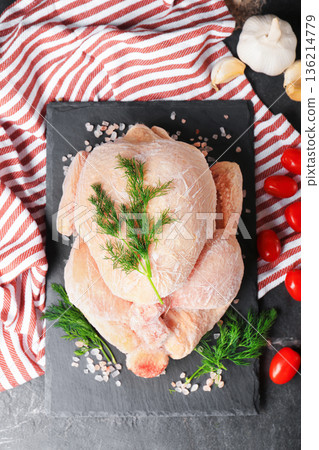 Top view of whole frozen chicken on black slate board with dill and tomatoes. Raw poultry meat covered in frost ready for cooking on dark table. High quality photo 136214779