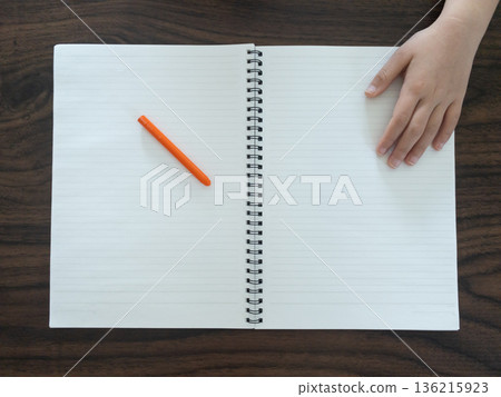 孩子的手放在木桌上的筆記本和橘色鋼筆旁邊(書頁之間有空隙)。 孩子的手放在木桌上的筆記本和橘色鋼筆旁邊(書頁之間有空隙)。 136215923