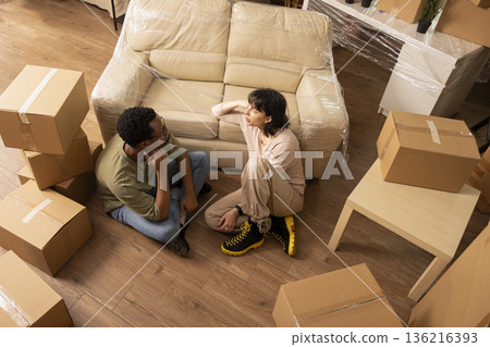 Young husband and wife sit on floor with moving boxes, talking and bonding together in cozy living room. Modern couple discussing future decorations and planning new home arrangements. Young husband and wife sit on floor with moving boxes, talking and bonding together in cozy living room. Modern couple discussing future decorations and planning new home arrangements. 136216393
