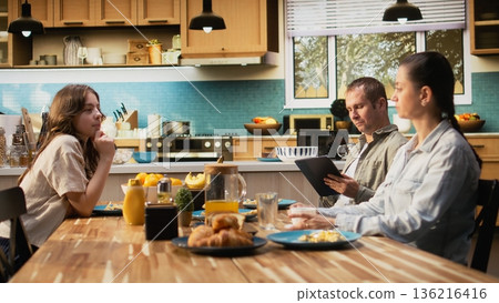 Working dad on tablet ignoring daughter and family at breakfast table, absorbed disconnected man distracted by his remote work. Inattentive parent distant with his girl and wife. 136216416