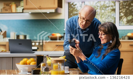 Senior woman and her husband laughing and checking smartphone at the breakfast table to enjoy a cozy meal with toast and coffee. Chatting and reflecting warm retirement lifestyle. Camera B. Senior woman and her husband laughing and checking smartphone at the breakfast table to enjoy a cozy meal with toast and coffee. Chatting and reflecting warm retirement lifestyle. Camera B. 136216428