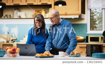 Aged husband and wife browsing laptop apps at counter in home kitchen, enjoying coffee and peaceful relaxation. Joyful lifestyle, satisfied wellbeing and relaxed moments in retirement. Camera B. Aged husband and wife browsing laptop apps at counter in home kitchen, enjoying coffee and peaceful relaxation. Joyful lifestyle, satisfied wellbeing and relaxed moments in retirement. Camera B. 136216441