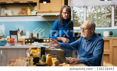 Mature old man browsing internet on laptop in a calm kitchen moment with coffee cup, highlighting relaxed lifestyle in retirement. Peaceful wellbeing and domestic life indoors. Camera B. Mature old man browsing internet on laptop in a calm kitchen moment with coffee cup, highlighting relaxed lifestyle in retirement. Peaceful wellbeing and domestic life indoors. Camera B. 136216443