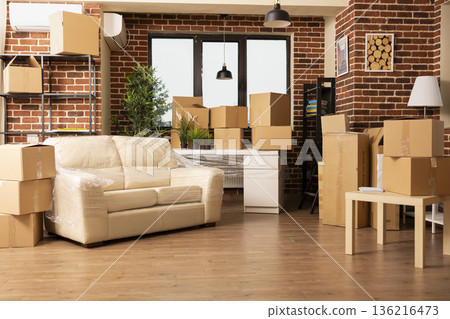Vacant apartment living room with stacked cardboard boxes and furniture arranged near brick wall. Packed items awaiting movers as people prepare to move in or out of modern space. Vacant apartment living room with stacked cardboard boxes and furniture arranged near brick wall. Packed items awaiting movers as people prepare to move in or out of modern space. 136216473