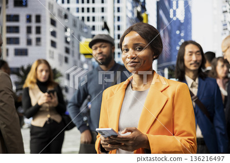 Portrait of african american businesswoman checking phone waiting at red light, walking the busy business district with a big urban crowd. Pedestrians group with different routines. Portrait of african american businesswoman checking phone waiting at red light, walking the busy business district with a big urban crowd. Pedestrians group with different routines. 136216497
