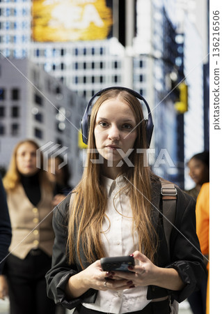 Portrait of young adult from gen z walking the streets with headset, stylish cool student waiting at a red light. Pedestrians of different ages on a busy avenue downtown, young girl. Portrait of young adult from gen z walking the streets with headset, stylish cool student waiting at a red light. Pedestrians of different ages on a busy avenue downtown, young girl. 136216506