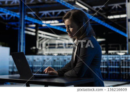 Data center female programmer using laptop to inspect infrastructure, updating network systems. IT professional using notebook to ensure stable server hub operations, overseeing rigs Data center female programmer using laptop to inspect infrastructure, updating network systems. IT professional using notebook to ensure stable server hub operations, overseeing rigs 136216609