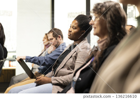 Black woman listening to presentation pitch with diverse people in attendance, business conference event with speaker. Chairs fill auditorium as participants attend professional summit. Black woman listening to presentation pitch with diverse people in attendance, business conference event with speaker. Chairs fill auditorium as participants attend professional summit. 136216610