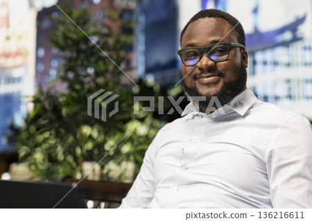 Portrait of african american person typing on laptop being seated on bench, blending relaxed lifestyle and business focus with the vibrant architecture. Busy city street within metropolitan area. 136216611