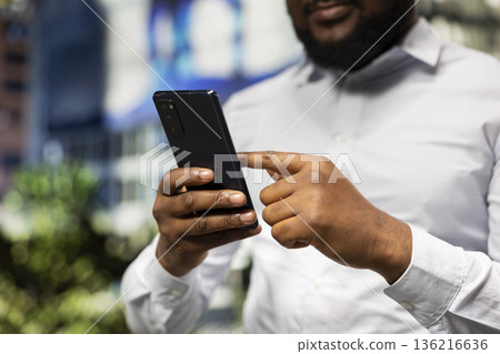 Black man navigating on mobile phone apps and walking the busy metropolitan street, urban roadway downtown with skyscrapers. Confident attitude reflecting a dynamic vibrant city life. 136216636