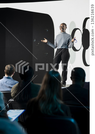 African american IT engineer introducing new mobile device launch with modern interface and design, marketing announcement. Mixed age participants group attend phone reveal in auditorium. 136216639