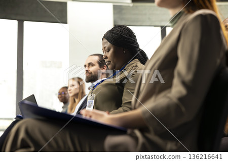 African american female attendee writes ideas on laptop at conference, business forum event with panel discussion and speaker. Multiracial audience takes notes for entrepreneurship. 136216641