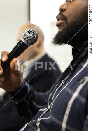 Close up of african american participant asking questions on microphone during panel discussion, engaging in debate at international summit. Business conference with presentation on stage. Close up of african american participant asking questions on microphone during panel discussion, engaging in debate at international summit. Business conference with presentation on stage. 136216643