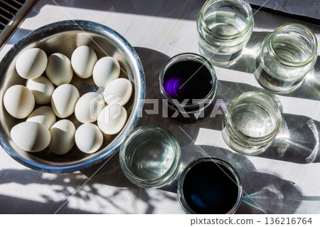 Top view of a bowl of white eggs with several glass jars of water and dark food dye solutions on a sunlit table Top view of a bowl of white eggs with several glass jars of water and dark food dye solutions on a sunlit table 136216764