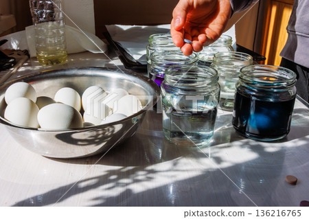 Hand holding a food dye tablet above a glass jar of water, preparing the eggs in a bowl nearby for easter egg coloring. 136216765