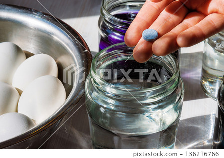 Hand holding a food dye tablet above a glass jar of water, with eggs in a bowl nearby for easter egg coloring. 136216766