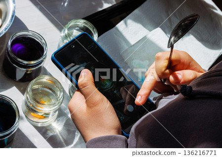 Top view of a hands using smartphone beside dye jars for easter egg coloring in sunlight Top view of a hands using smartphone beside dye jars for easter egg coloring in sunlight 136216771
