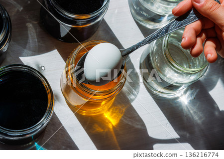 Top view of a white egg held on a spoon above a jar of yellow food dye, ready to dip, surrounded by clear water jars in bright sunlight. 136216774
