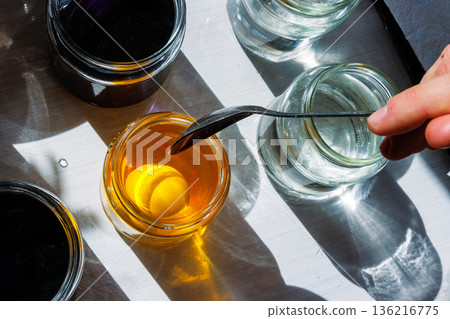 Top view of a white eggdipped into a jar of yellow food dye, surrounded by clear water jars in bright sunlight. Top view of a white eggdipped into a jar of yellow food dye, surrounded by clear water jars in bright sunlight. 136216775