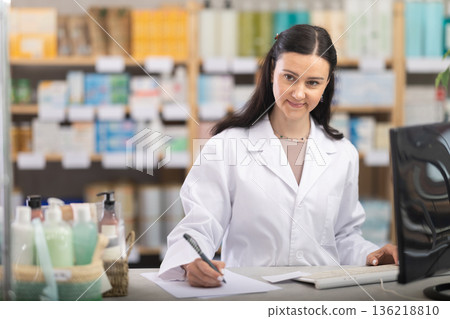 Woman pharmacy employee works with computer and notebook dairy. Woman pharmacy employee works with computer and notebook dairy. 136218810