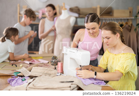 Teenage girl works on sewing machine while teacher and children learn how to cut fabric 136219024