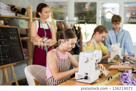 Teenage girl works on sewing machine while teacher and children learn how to cut fabric Teenage girl works on sewing machine while teacher and children learn how to cut fabric 136219072