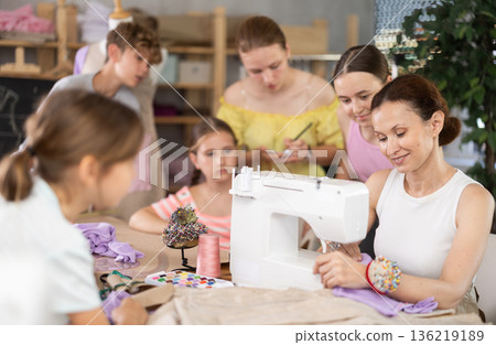 Woman teacher demonstrates how to sew on a sewing machine. Children gather around and watch 136219189
