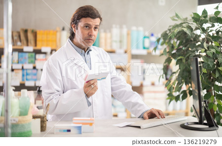 Friendly male pharmacist standing at drugstore with medical product in hands Friendly male pharmacist standing at drugstore with medical product in hands 136219279