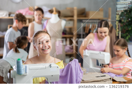 Teenage girl works on sewing machine while teacher and children learn how to cut fabric Teenage girl works on sewing machine while teacher and children learn how to cut fabric 136219376