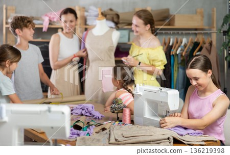 Teenage girl works on sewing machine while teacher and children learn how to cut fabric 136219398