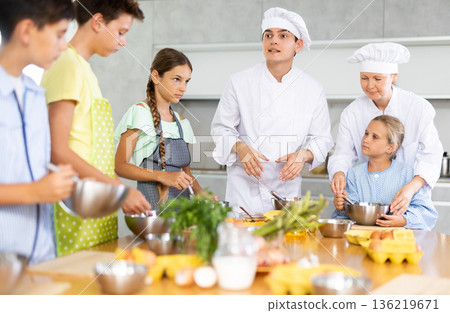 During cooking lesson, male cook tells children about culinary rules, woman assistant helps child During cooking lesson, male cook tells children about culinary rules, woman assistant helps child 136219671