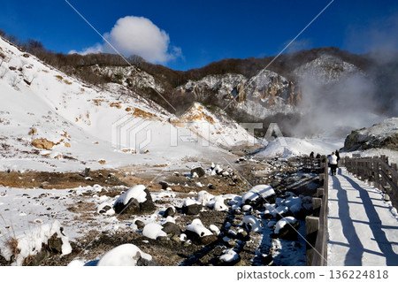 Noboribetsu Jigokudani on a clear winter day. A walking trail stretches through the snow and brown mountainside into the center of the valley. Noboribetsu Jigokudani on a clear winter day. A walking trail stretches through the snow and brown mountainside into the center of the valley. 136224818