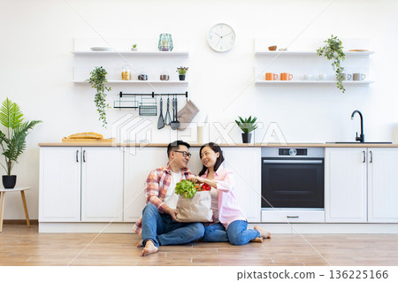 A happy couple sits on the floor of their modern kitchen, unpacking fresh produce from a paper bag A happy couple sits on the floor of their modern kitchen, unpacking fresh produce from a paper bag 136225166