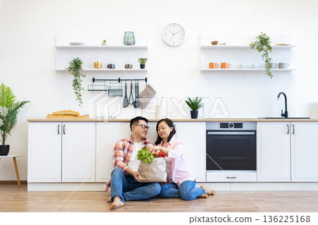 An Asian couple sits on the floor of their modern kitchen, unpacking fresh groceries together 136225168