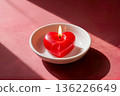 Red heart shaped burning candle in white saucer on red background 136226649