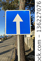 Blue rectangular one way arrow sign directs vehicles along a seaside street, captured on a bright sunny day.  136227076