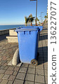 A blue plastic garbage bin stands on a seaside promenade under a clear sky, placed next to a curved concrete stopper.  136227077